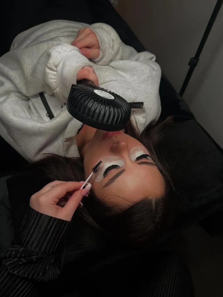 Especialista de belleza aplicando extensiones de pestañas a una cliente mientras usa un mini ventilador negro.