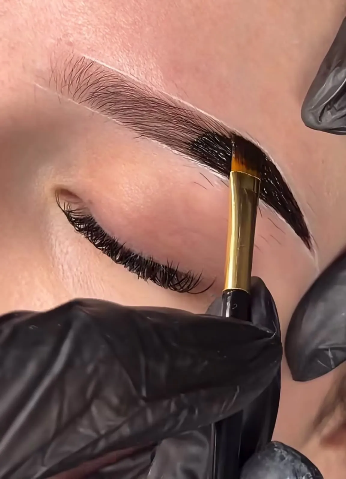 Especialista aplicando tinte o henna oscura en las cejas con un pincel plano dorado.